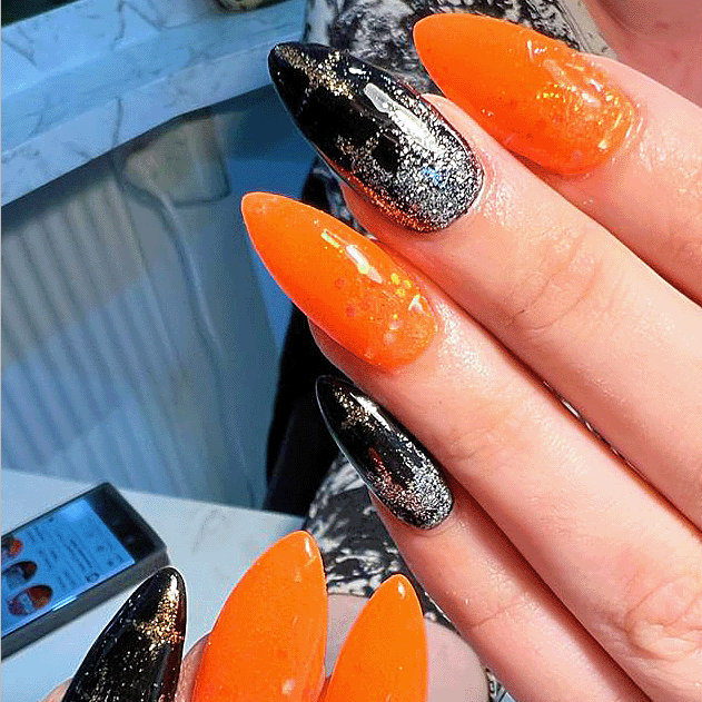Hand with long almond-shaped nails painted in glossy black with silver glitter and vibrant orange polish with subtle shimmer.