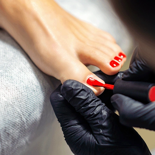 Close-up of a pedicure with red nail polish being applied to toenails by a person wearing black gloves.